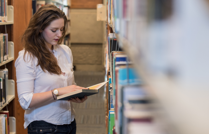 young woman standing between two shelves reading book