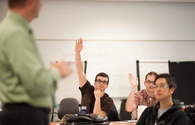 Students in a classroom, one raising hand to ask a question