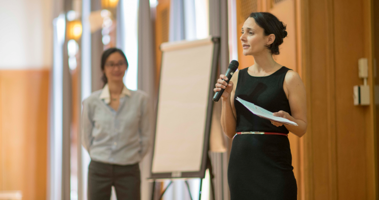 Person holding microphone presenting with second person in background