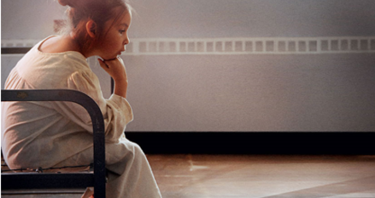 A young Indigenous girl sits on a chair in an institutional setting, hand on fist, looking at the floor. The shadow of a cross appears on the floor.