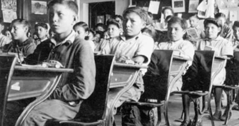 Historical image of young Indigenous children sitting in a classroom looking miserable.