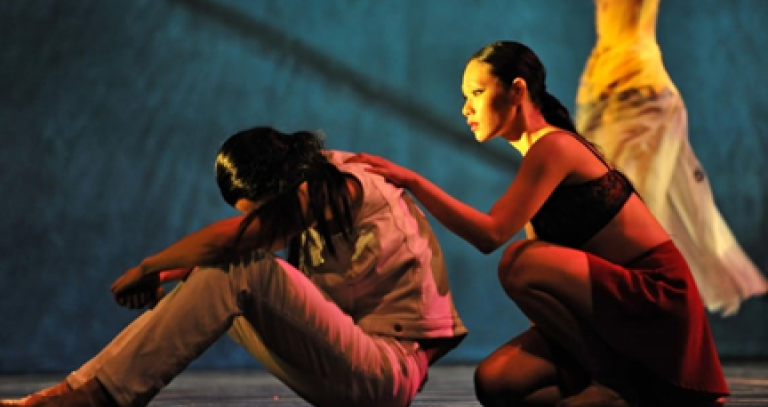 video still from a dance: a woman sits with her head in her arms and knees pulled up, while another woman kneels behind her and touches her back