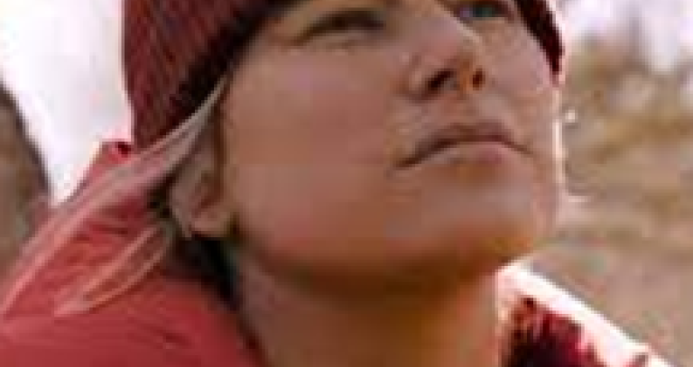 An Indigenous person in a hat and jacket stands by a tree and looks confidently off-camera.